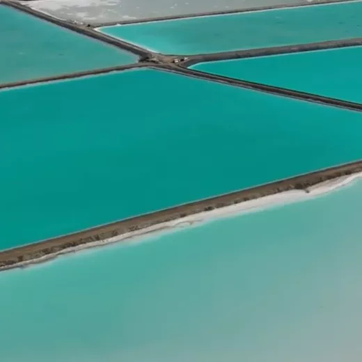 Aerial view of salt evaporation ponds divided into sections by narrow pathways, with vibrant shades of turquoise and green water.