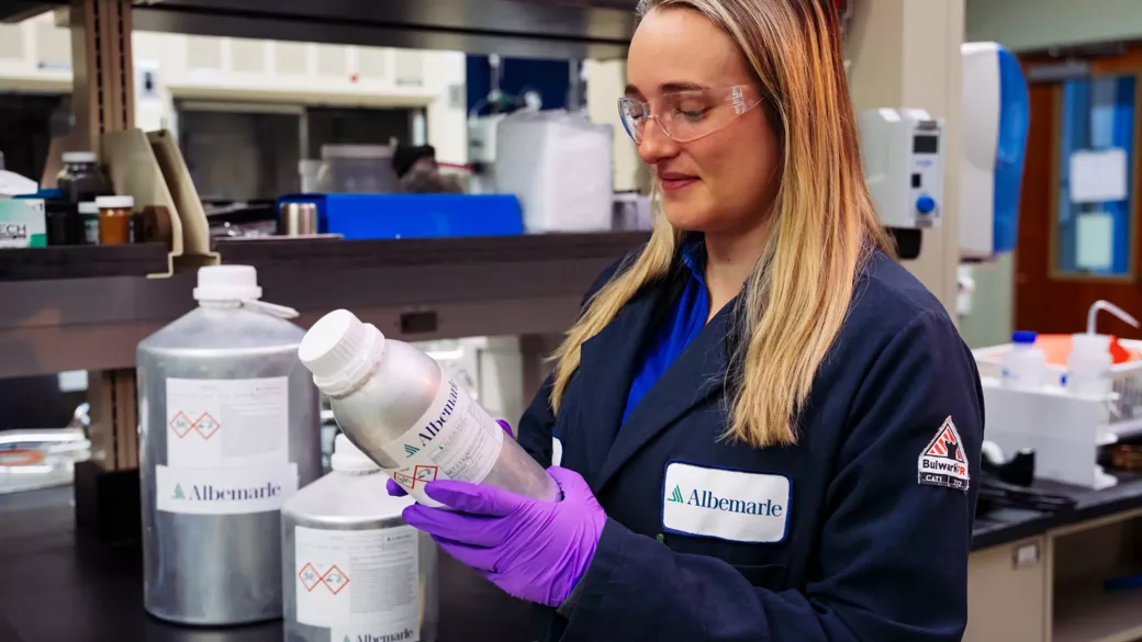 An Albemarle employee in a laboratory wearing safety goggles and purple gloves, holding a silver bottle with a label. They are dressed in a dark lab coat with a company logo. Shelves with various lab equipment and containers are visible in the background.