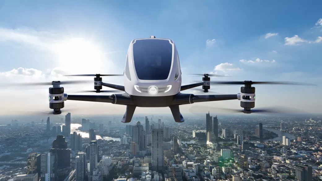 A futuristic drone or flying taxi with four rotors, featuring a sleek white cabin, hovers above a cityscape under a clear blue sky.