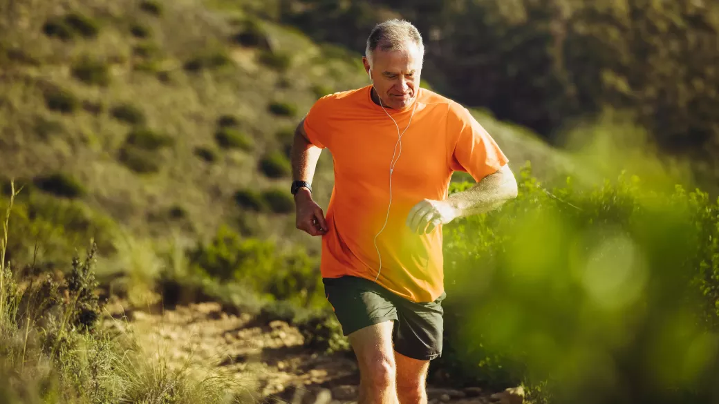 A middle-aged man in an orange t-shirt and black shorts jogging on a trail, with greenery around. He is wearing headphones and checking a watch on his left wrist.