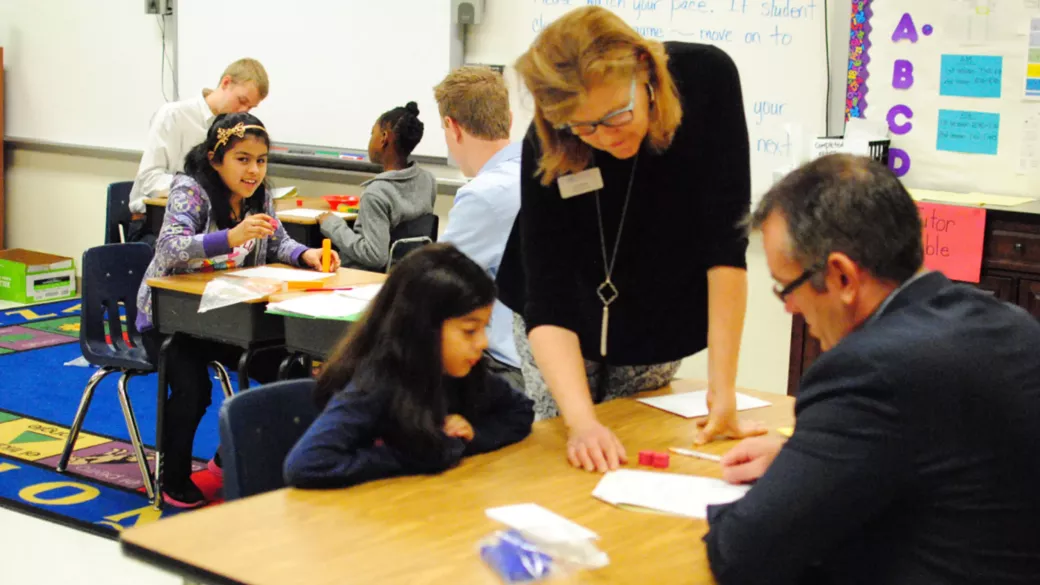 An elementary school classroom with a diverse group of students engaged in learning. A female teacher assists a young girl at a table, while other students work with adults in the background.