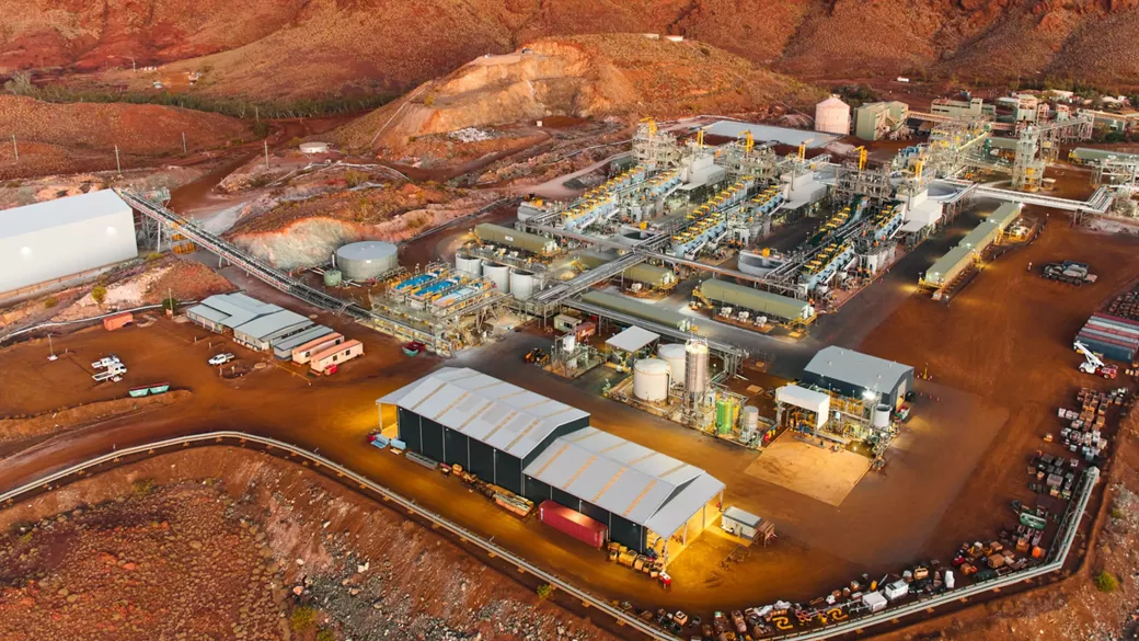 Aerial view of a large industrial complex set against a rugged, arid landscape. The facility features multiple buildings, structures, and machinery, with roads connecting different sections. Various equipment, vehicles, and storage tanks are visible throughout the site.