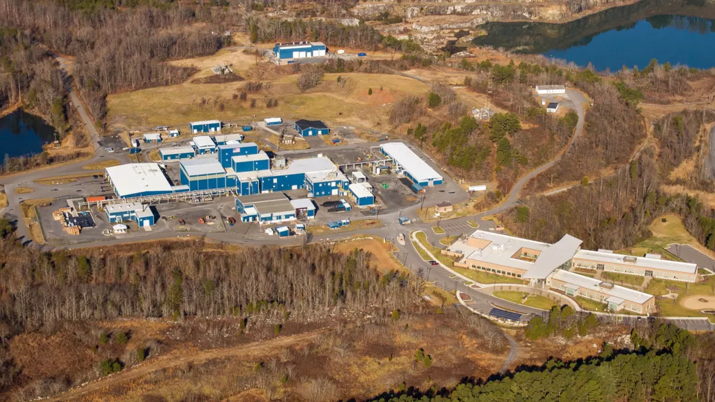 Aerial view of a large industrial facility with multiple blue-roofed buildings surrounded by trees and greenery. There is a smaller complex with white-roofed structures nearby. The area features paved roads, parking spaces, and two bodies of water in the background.