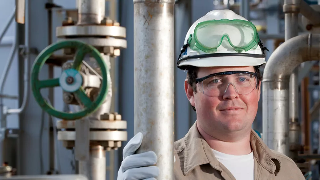 A worker wearing a hard hat with goggles and safety gloves, smiling at the camera in an industrial setting with metal pipes and a large valve in the background.