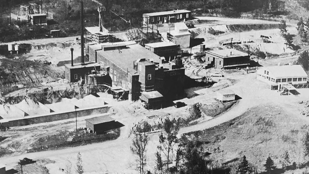 Historic black and white photo of an early 20th-century mining operation with various buildings, equipment, and earth mounds in a forested area.