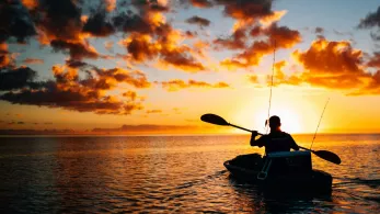 A person in a kayak fishing at sea, silhouetted against a vibrant sunset with golden clouds.