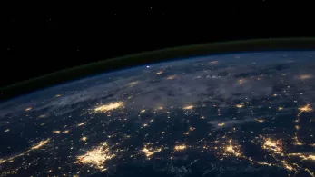 A view of earth from space at night showing the planet's curved horizon. Some clouds are illuminated by city lights. Clusters of city lights showing in certain areas looking like connected light streams.