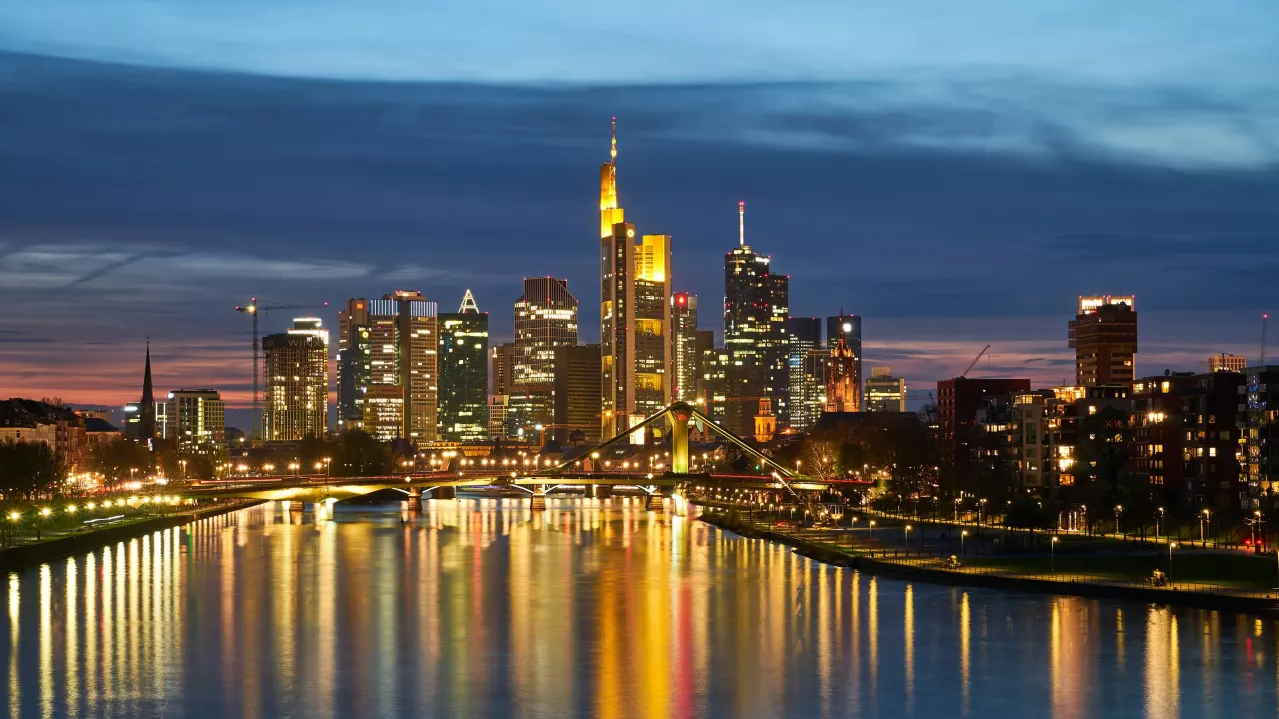 Night view of Frankfurt's skyline with illuminated skyscrapers reflecting in a river, featuring a bridge in the foreground and a sunset backdrop.