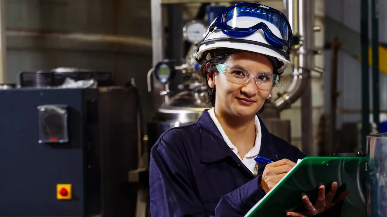 A female Albemarle employee wearing safety gear, including a hard hat, protective glasses, and ear protection, holds a pen and green clipboard while standing in an industrial setting. Machinery and metal pipes can be seen in the background.