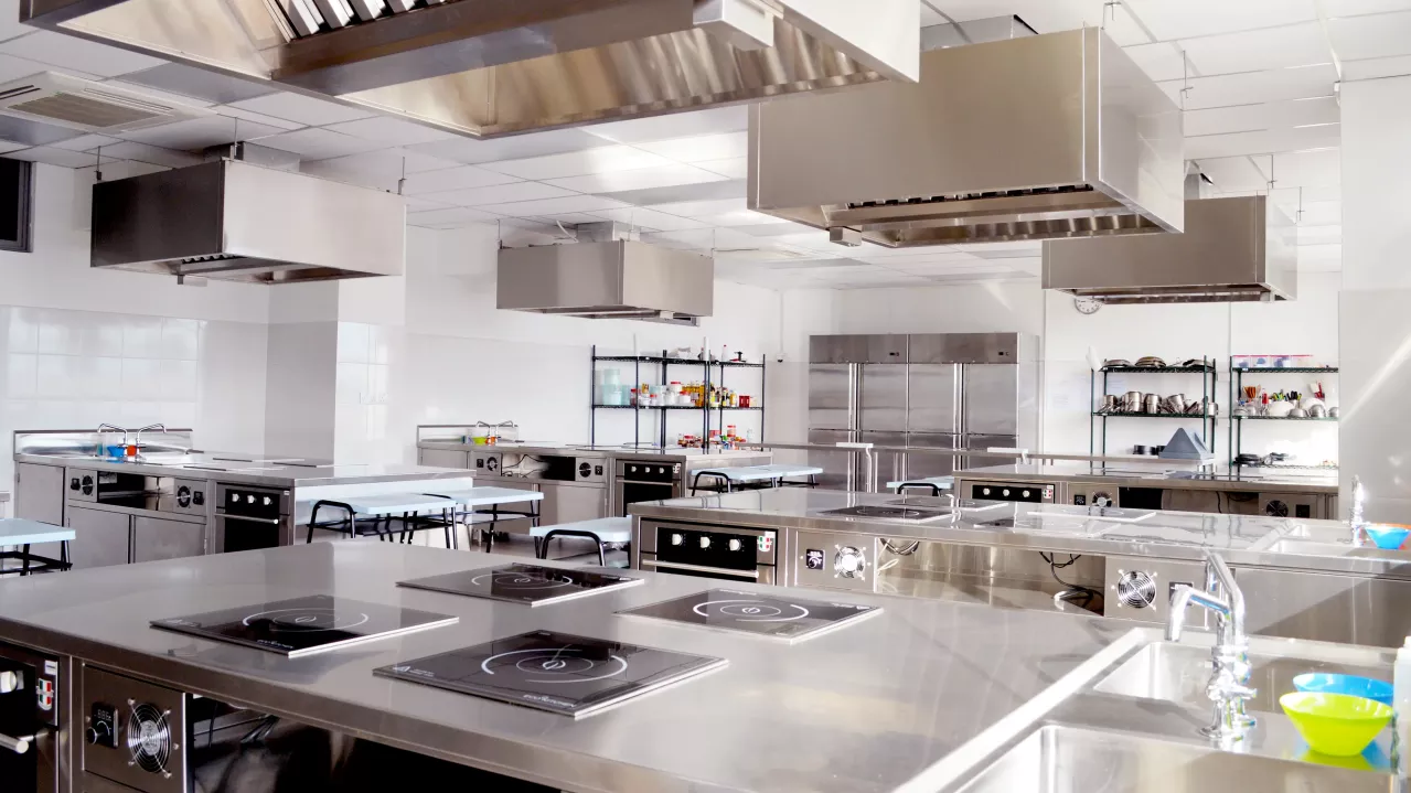A modern commercial kitchen with stainless steel countertops, multiple cooking stations with built-in electric stoves, overhead exhaust hoods, and scattered utensils. Brightly lit and spacious.