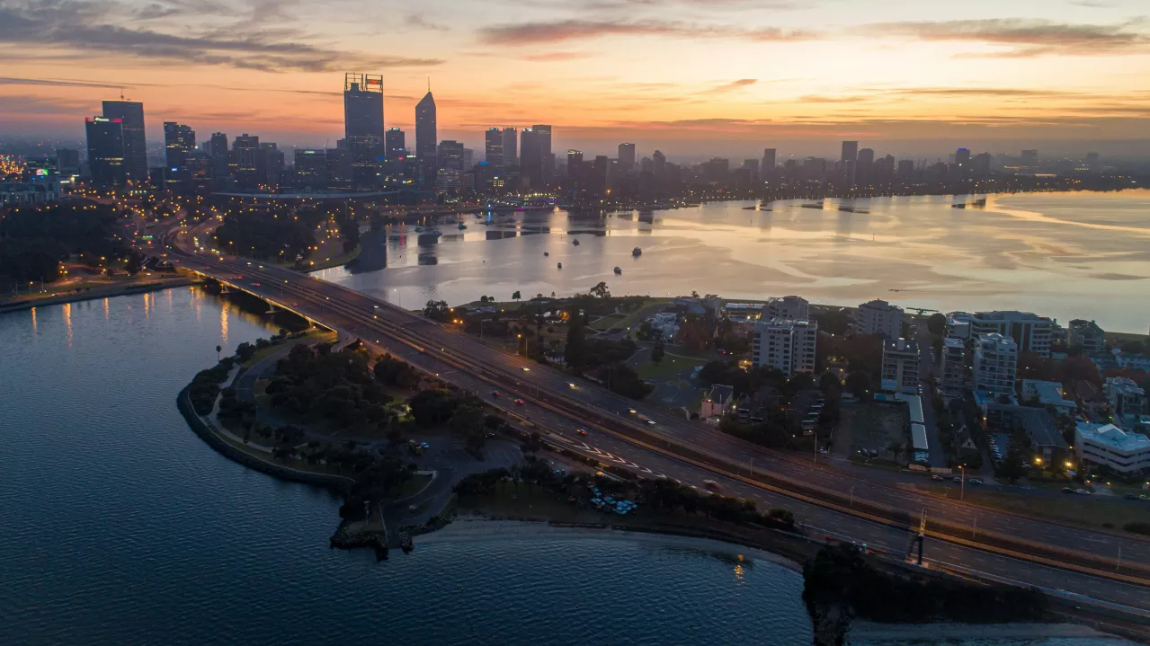 Aerial view of Perth's skyline at sunset, with the Swan River running through it, reflecting the sky’s colors, and a multi-lane highway bordering the waterfront