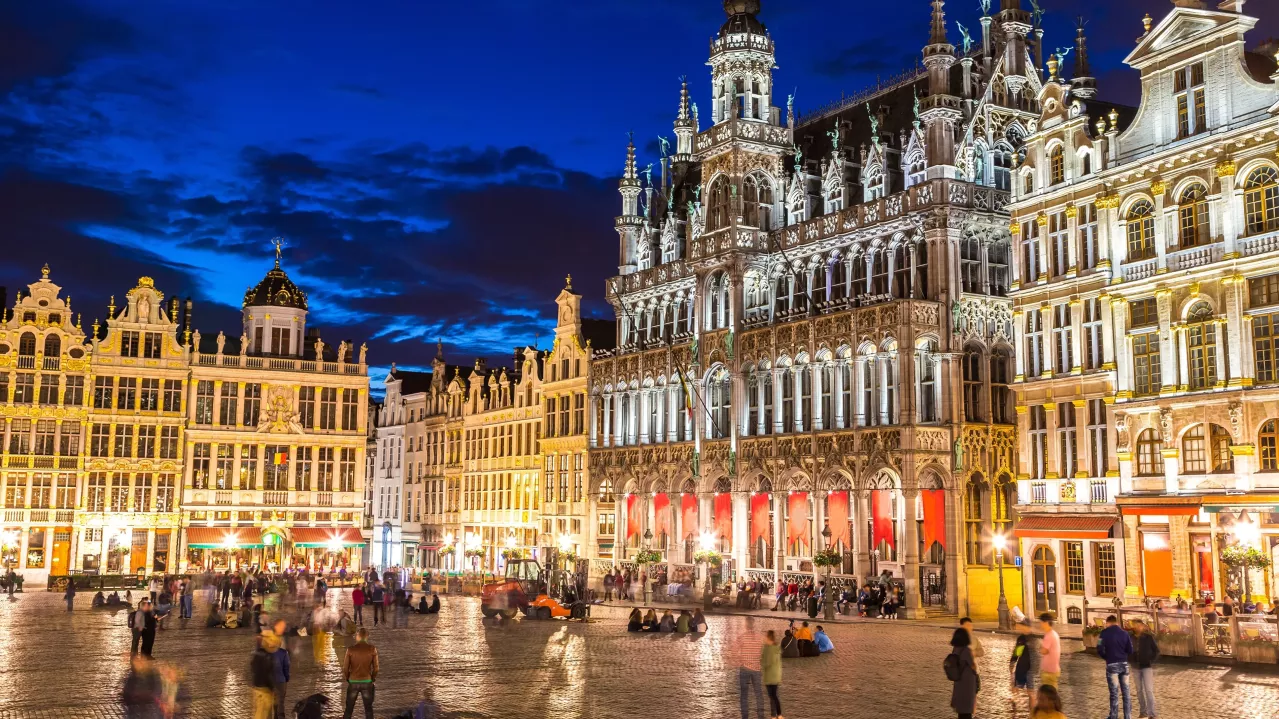 The Grand Place in Brussels illuminated at night, featuring ornate, historic buildings and a lively crowd.