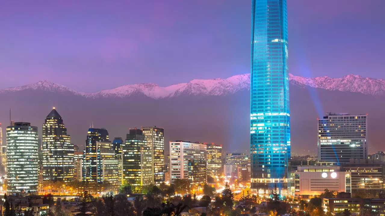 Nighttime Santiago skyline featuring a tall illuminated skyscraper center right, surrounded by numerous buildings, with snow-capped mountains in the background, under a purple sky.