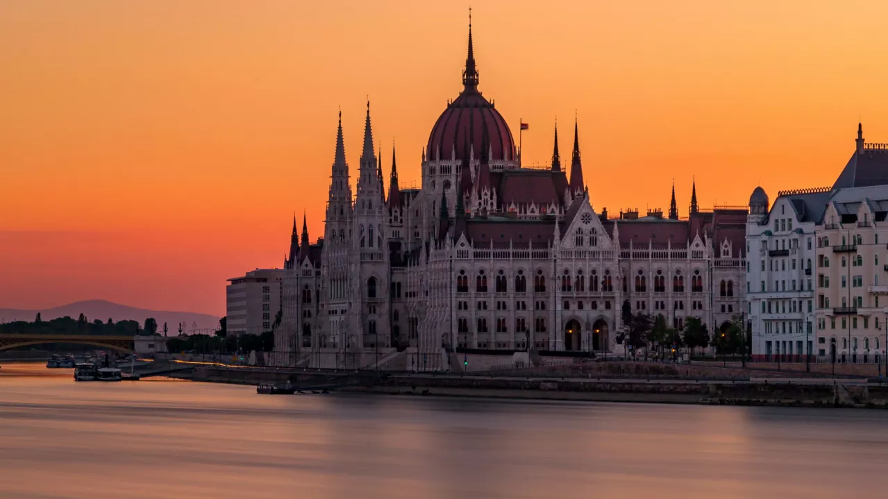 A panoramic view of the Hungarian Parliament Building in Budapest at sunset, with the Danube River in the foreground and a vibrant orange sky.