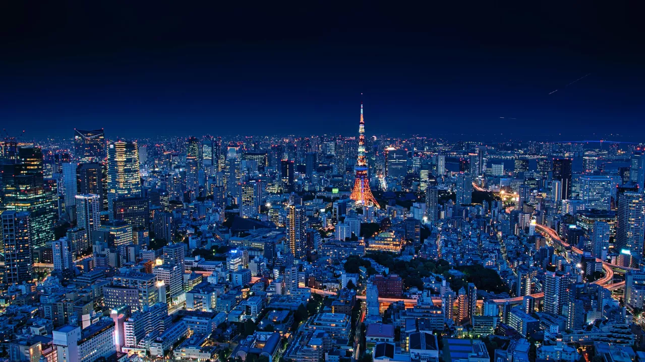 Aerial view of Tokyo at night, brightly illuminated with dense skyscrapers and a prominent tower glowing orange in the center, under a dark blue sky.