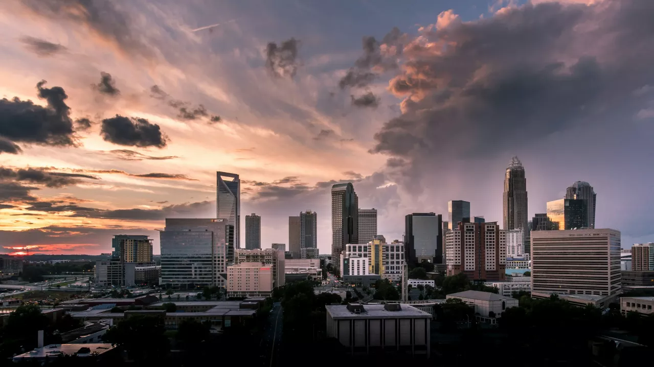 Skyline of Charlotte North Carolina at dusk, featuring a mix of high-rise buildings under a dramatic cloudy sky with a hint of sunset on the horizon.