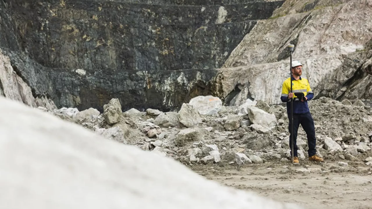 A lone worker in a yellow and blue uniform stands on rocky terrain at a quarry site, holding a surveying pole. The background features a steep rock face, with various shades of gray and brown indicating the layers of earth and mineral deposits.