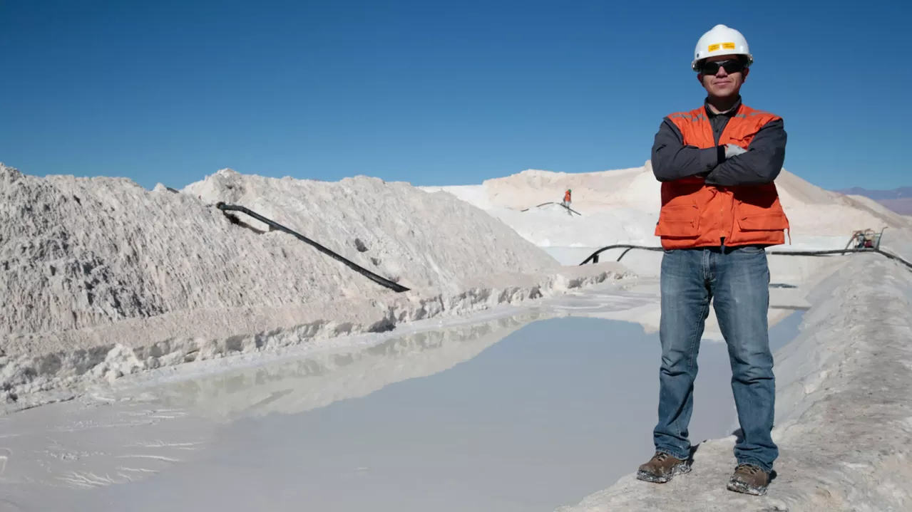 A man in a hard hat and orange vest stands with crossed arms at a salt mine, with large white salt piles and machinery in the background under a clear blue sky.