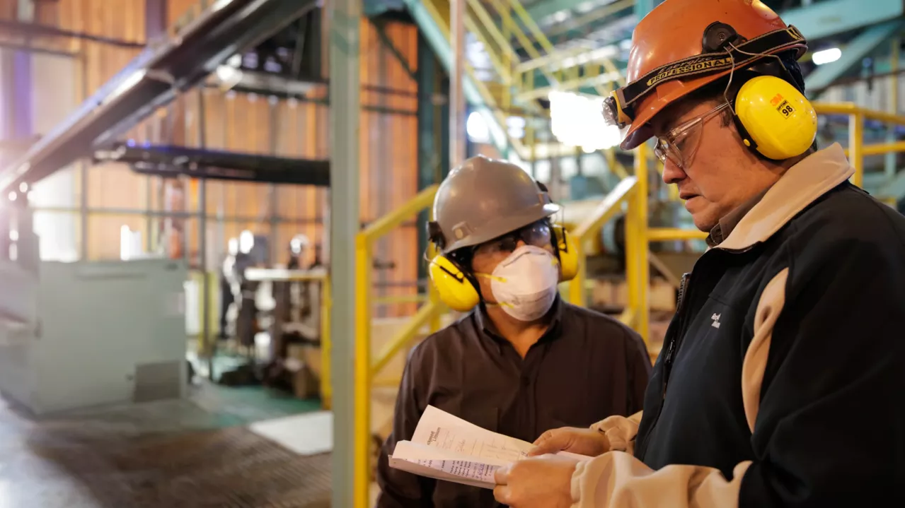 Two industrial workers are in conversation within a factory setting. Both are wearing hard hats and ear protection; one also wears a face mask. The individual on the right holds and refers to a document. Yellow railings and industrial machinery are visible in the background.