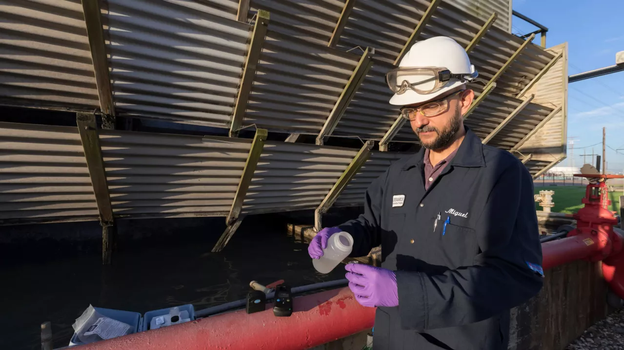 A man wearing a hard hat, safety glasses, and purple gloves is examining a water sample at an industrial facility. He is standing next to large metal structures and valves.