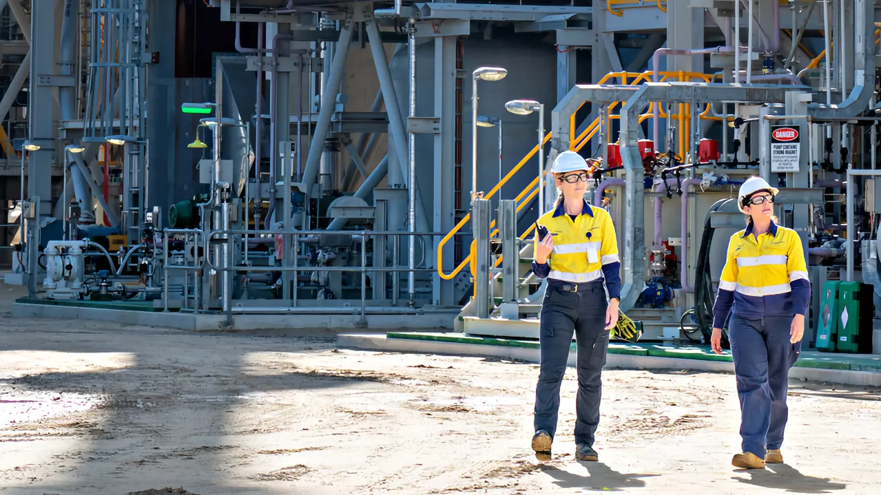 Two individuals wearing yellow safety jackets and white hard hats walk through an industrial facility. They are surrounded by machinery and pipelines. The weather appears sunny, and shadows fall on the ground, indicating bright sunlight.