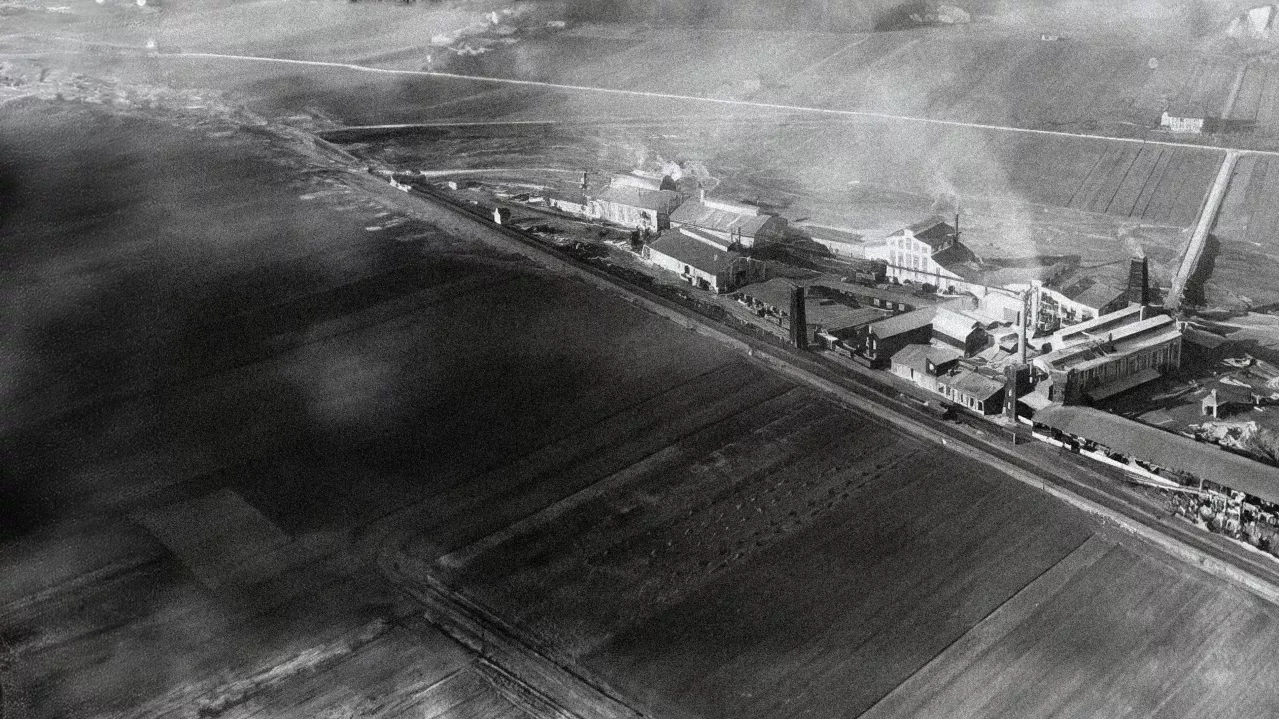 Aerial black and white photo of an industrial complex with surrounding fields, featuring distinct buildings, smokestacks, and a large expanse of open land.
