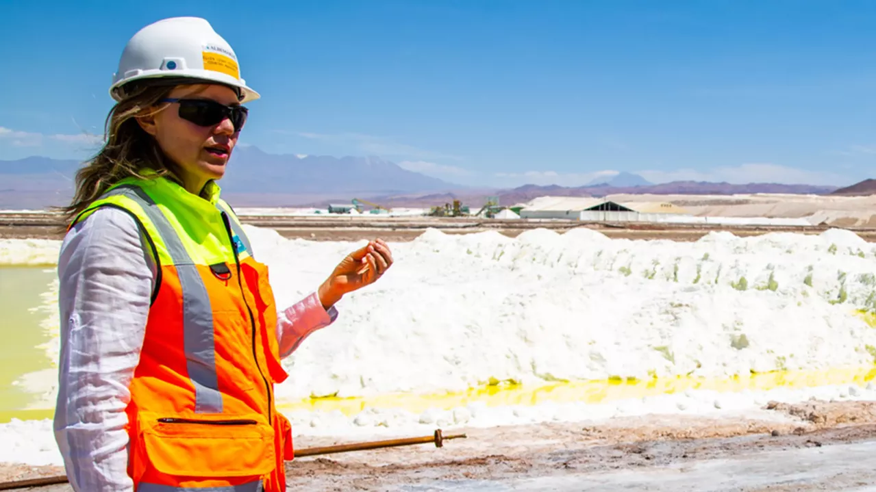 A person wearing a white hard hat, sunglasses, and an orange safety vest stands outdoors by a large salt flat under a clear blue sky. The person appears to be gesturing with their hand while discussing something. Industrial buildings and mountains are visible in the background.