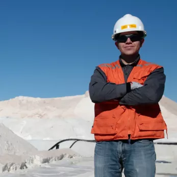A man in a hard hat and orange vest stands with crossed arms at a salt mine, with large white salt piles and machinery in the background under a clear blue sky.