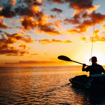 A person in a kayak fishing at sea, silhouetted against a vibrant sunset with golden clouds.