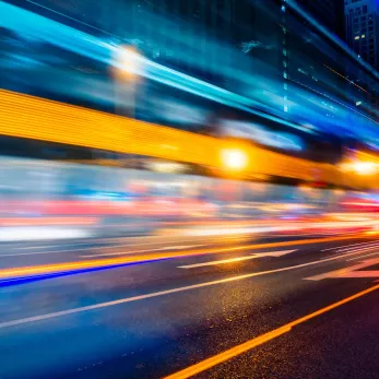 Long exposure shot of a busy city street at night showing streaks of lights from moving vehicles, with buildings illuminated by streetlights and signs.