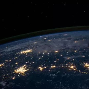 A view of earth from space at night showing the planet's curved horizon. Some clouds are illuminated by city lights. Clusters of city lights showing in certain areas looking like connected light streams.
