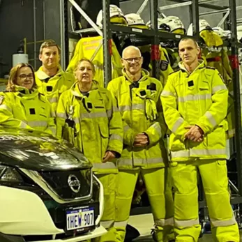 Five firefighters wearing high-visibility yellow gear stand together in a garage. Behind them are shelves with helmets and other firefighting equipment. A car with an Australian license plate is partially visible in the foreground.