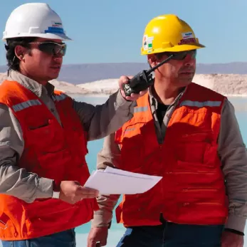 Two men wearing white and yellow hard hats and orange safety vests stand outdoors. One man holds a radio while the other holds a clipboard. They both wear sunglasses and are looking into the distance, with a body of water and mountains in the background.