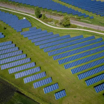 Aerial view of a large solar power plant with rows of blue solar panels arranged in a grid pattern on a vast green field. A pathway and some trees can be seen in the background near the top of the image.