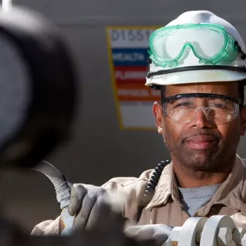 A worker wearing a white hard hat, safety goggles, and gloves operates equipment in an industrial setting. He stands behind machinery, maintaining a focused expression, with other industrial components and a gauge partially visible in the foreground.