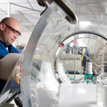 A person wearing safety glasses and a blue lab coat is working in a laboratory. They are using a glove box with a transparent dome to handle materials inside. Laboratory equipment and a large clear container are visible in the background.