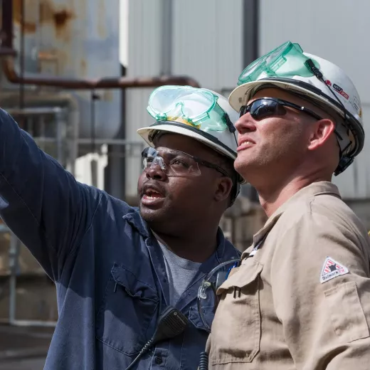 Two diverse industrial workers wearing protective gear, including helmets, safety glasses, and earmuffs, are engaged in a discussion at a worksite. One worker is pointing towards something off-camera while both look attentively in the same direction.
