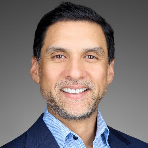 Headshot of Neil Sheorey, Executive Vice President and Chief Financial Officer who is smiling at the camera, showcasing short black hair, slight stubble, and wearing a dark suit with a light blue shirt.. 