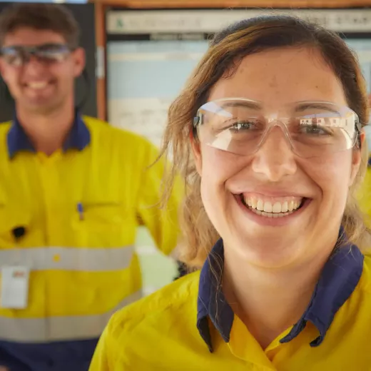 Two workers in yellow reflective safety uniforms and clear safety glasses smile at the camera. The woman in the foreground is in sharp focus, while the man in the background is slightly blurry. They are in what appears to be an industrial or construction setting.