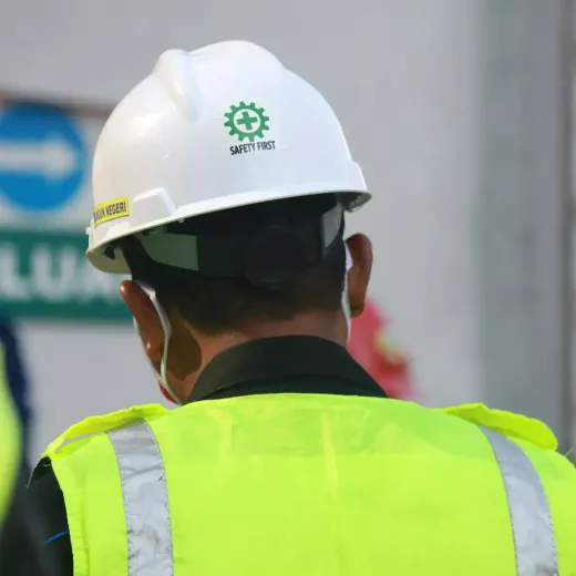 A construction worker viewed from behind wearing a white helmet labeled "safety first" and a high-visibility yellow vest, standing at a work site.
