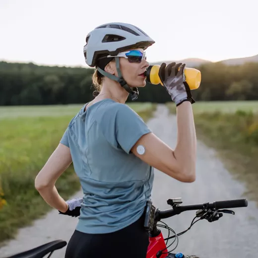 A woman in cycling gear drinking from a yellow water bottle while standing next to her bicycle on a dirt trail, with a serene natural landscape in the background. She wears sunglasses and a helmet.