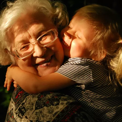 A joyful elderly woman with glasses embracing a laughing young child in sunlight, sharing a tender moment.