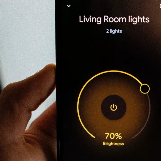 A close-up of a hand holding a smartphone displaying a smart home app interface for controlling living room lights, with a brightness setting at 70%.