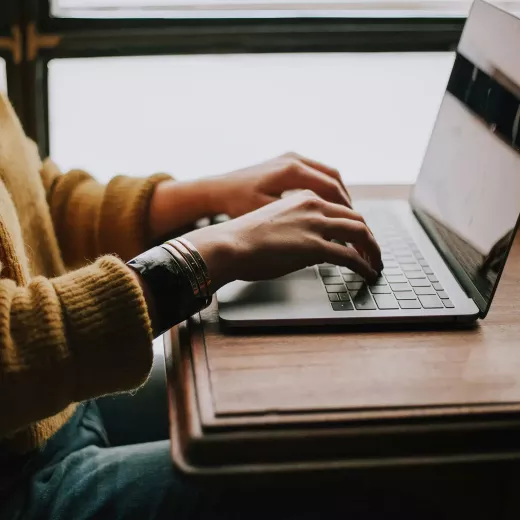 A person in a yellow sweater typing on a laptop with a strong, fire-resistant circuit board. The laptop is placed on a wooden desk, near a window with natural light streaming in.