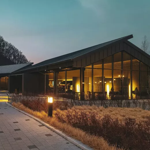 A modern office building with energy efficient strong windows illuminated in the evening, surrounded by dried grass and a lit pathway leading to it.
