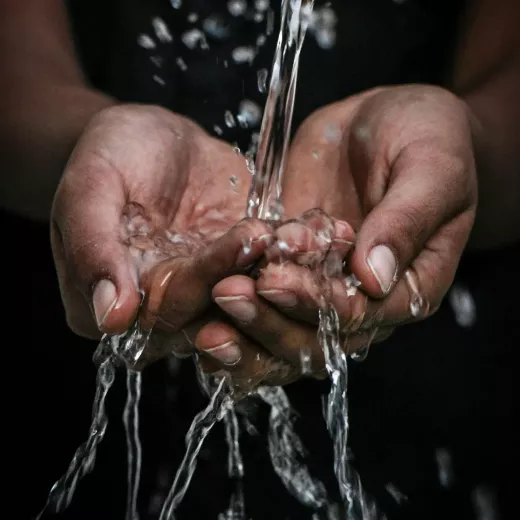 A close-up image of a person's cupped hands catching clear, splashing water, highlighting the motion of the water and the detail of the hands.