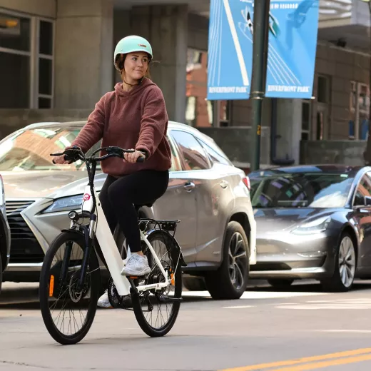 A woman wearing a helmet and casual clothing rides a white and red electric bike on a city street, with cars and city park banners in the background.