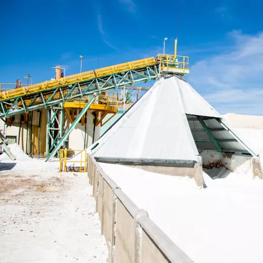 A yellow conveyor belt on a green framework is positioned above a large, metal-roofed conical structure, which appears to be for storing or processing bulk salt or sand. The surrounding area is covered in salt material, with a clear blue sky above.