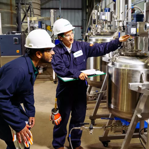 Two individuals in safety gear, including hard hats and glasses, examine equipment in an industrial setting. One person points to a device on a large stainless steel container, while holding a clipboard. They are surrounded by industrial machinery and stainless steel tanks.