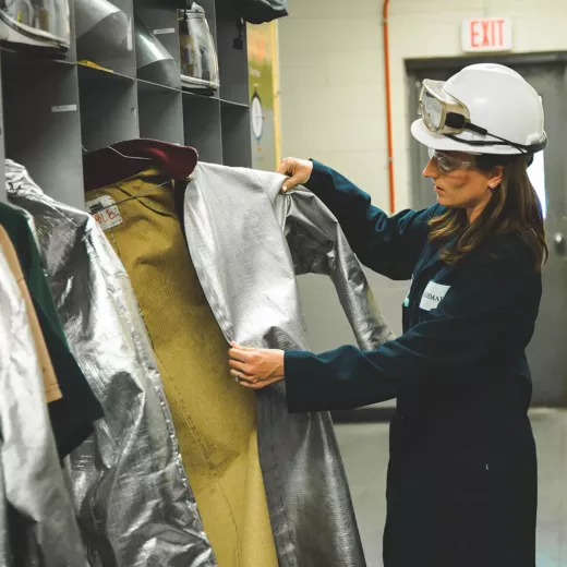 A woman wearing safety glasses and a hard hat examines a metallic, protective coat in a storage area, likely in an industrial or lab setting. She is holding the coat open, inspecting the inside lining, with helmets and other clothing hanging in the background.